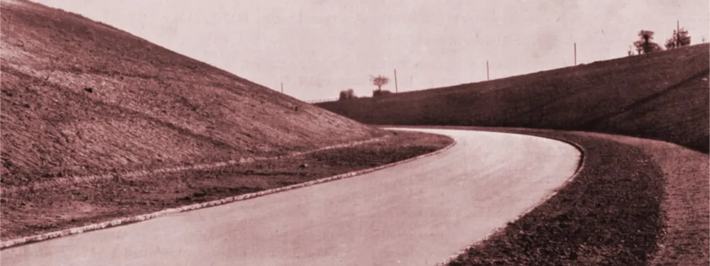 A black and white photograph showing a wide, brand new road, with no road markings, curving away through a deep man-made cutting.