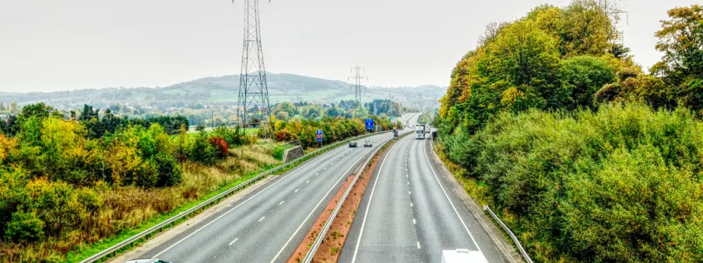 View of a modern dual carriageway that curves away into the distance; a line of pylons follows the route and dramatic hills are visible on the horizon