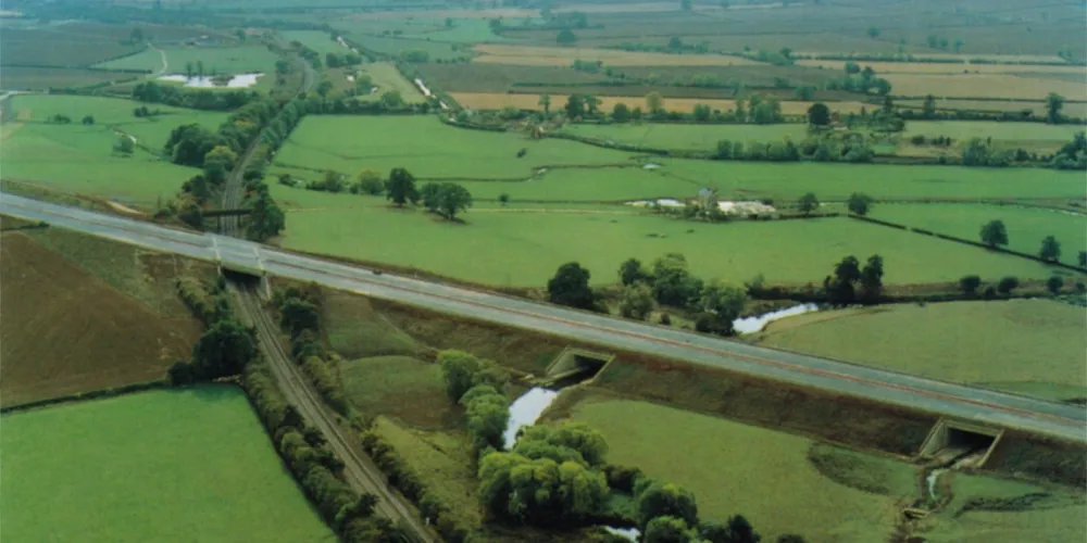 Aerial photograph of a new motorway crossing flat green countryside on an embankment. It bridges a canal and a railway, which run from the bottom of the picture towards the top, and there are patches of woodland between the green fields