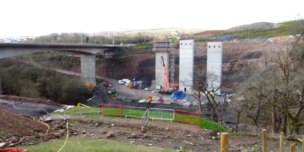 Photograph of a construction site in a valley. A long concrete road viaduct crosses the valley on the left; in the centre of the picture three tall concrete towers have been built to support three new viaducts alongside it.