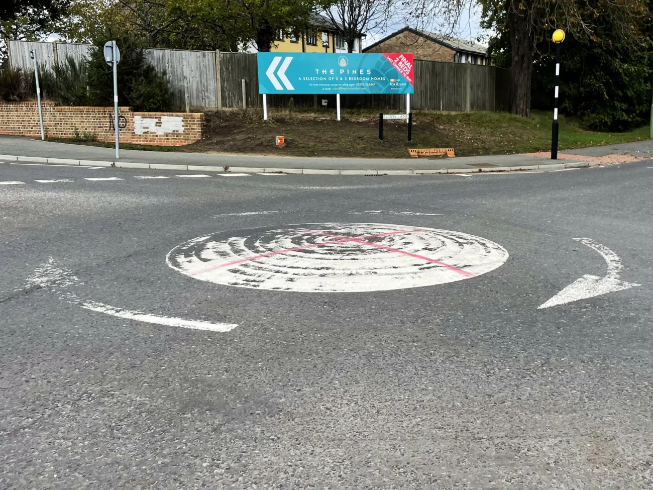 A mini-roundabout with worn white paint has two red lines spray-painted on to form a rudimentary St George's Cross
