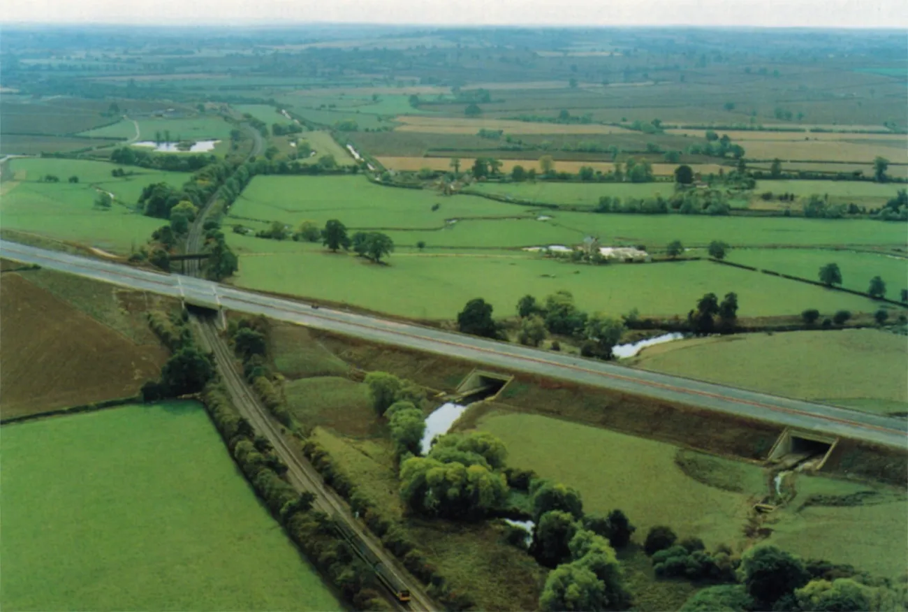 Aerial photograph of a new motorway crossing flat green countryside on an embankment. It bridges a canal and a railway, which run from the bottom of the picture towards the top, and there are patches of woodland between the green fields