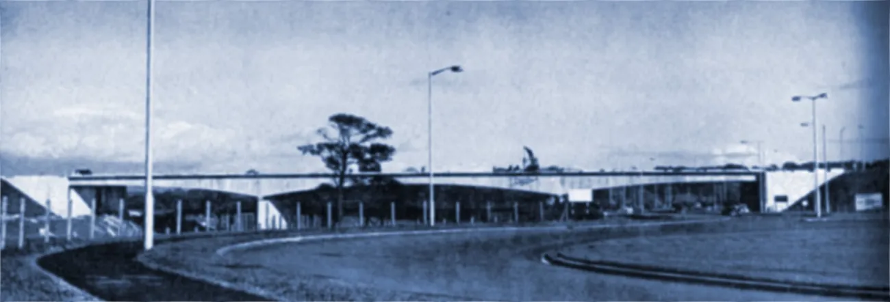 Black and white photograph of a roundabout forming part of a motorway interchange, with streetlights and trees in the foreground. A long arched motorway bridge stretches across the background from left to right
