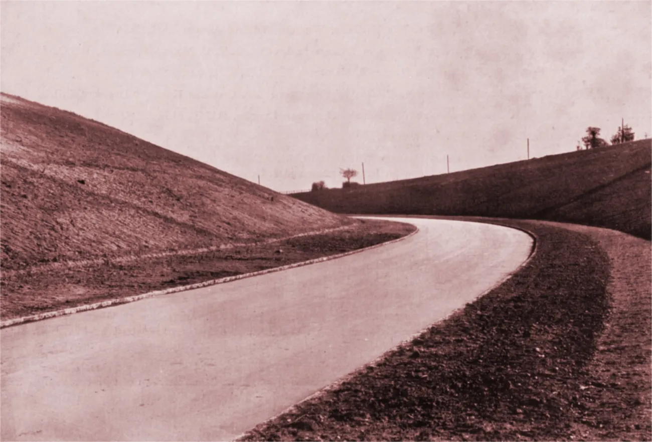 A black and white photograph showing a wide, brand new road, with no road markings, curving away through a deep man-made cutting.
