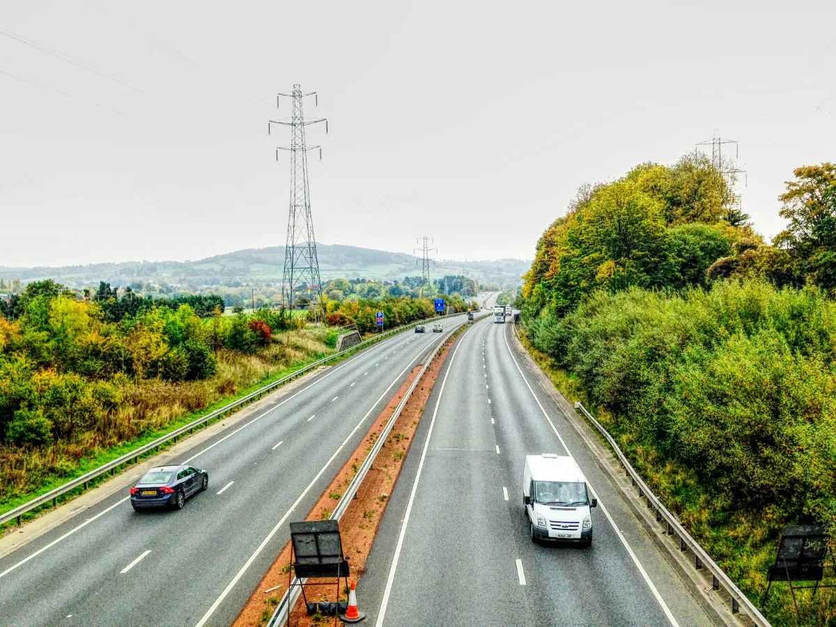View of a modern dual carriageway that curves away into the distance; a line of pylons follows the route and dramatic hills are visible on the horizon