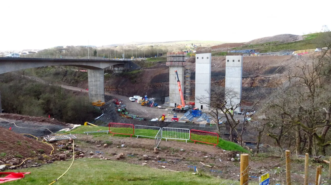 Photograph of a construction site in a valley. A long concrete road viaduct crosses the valley on the left; in the centre of the picture three tall concrete towers have been built to support three new viaducts alongside it.