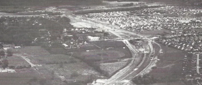Black and white aerial photograph of a dual carriageway under construction, curving between fields and housing estates