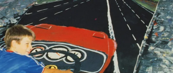 Photograph of a young boy sitting on a large painting of a car on a motorway, pretending to hold the steering wheel in the picture